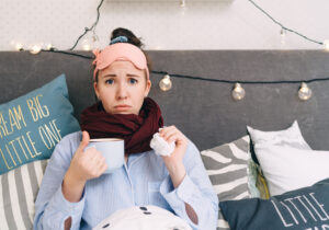 Young woman in bed, drinking tea and holding a tissue to sooth her cold.