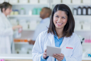 Beautiful mid adult Asian female pharmacist fills a prescription. She is holding a digital tablet. People are working in the background.