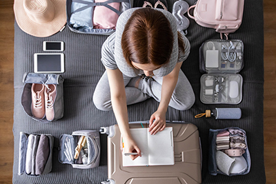 Smiling tourist woman packing suitcase to vacation writing paper list sitting on bed at home. Female getting ready to travel trip organization things storage in comfy cases bags use konmari method
